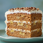 Close-up of a generous slice of moist hummingbird cake with cream cheese frosting and chopped pecans on a light blue-green plate.